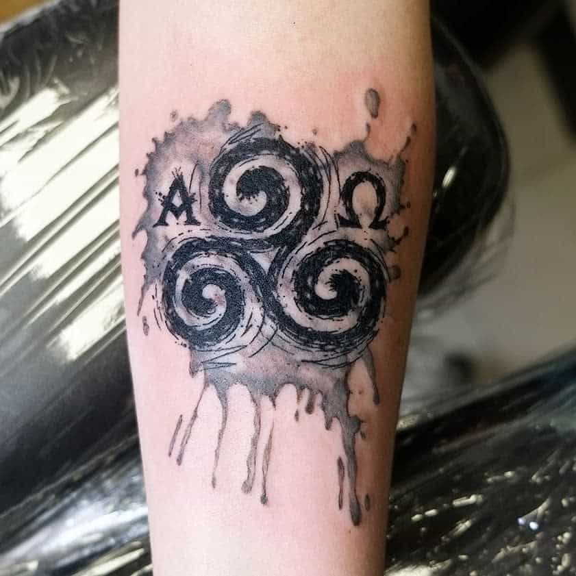 Abstract Scottish tattoo featuring a triskelion symbol with black watercolor splatter effects and alpha-omega symbols
