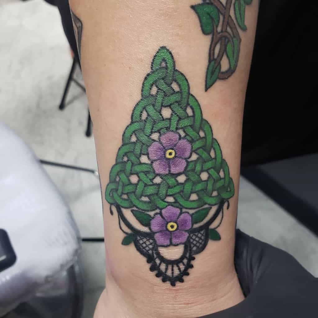 A green Scottish Celtic knot tattoo in a triangular shape, accented with two purple flowers and black lace-like detailing at the bottom