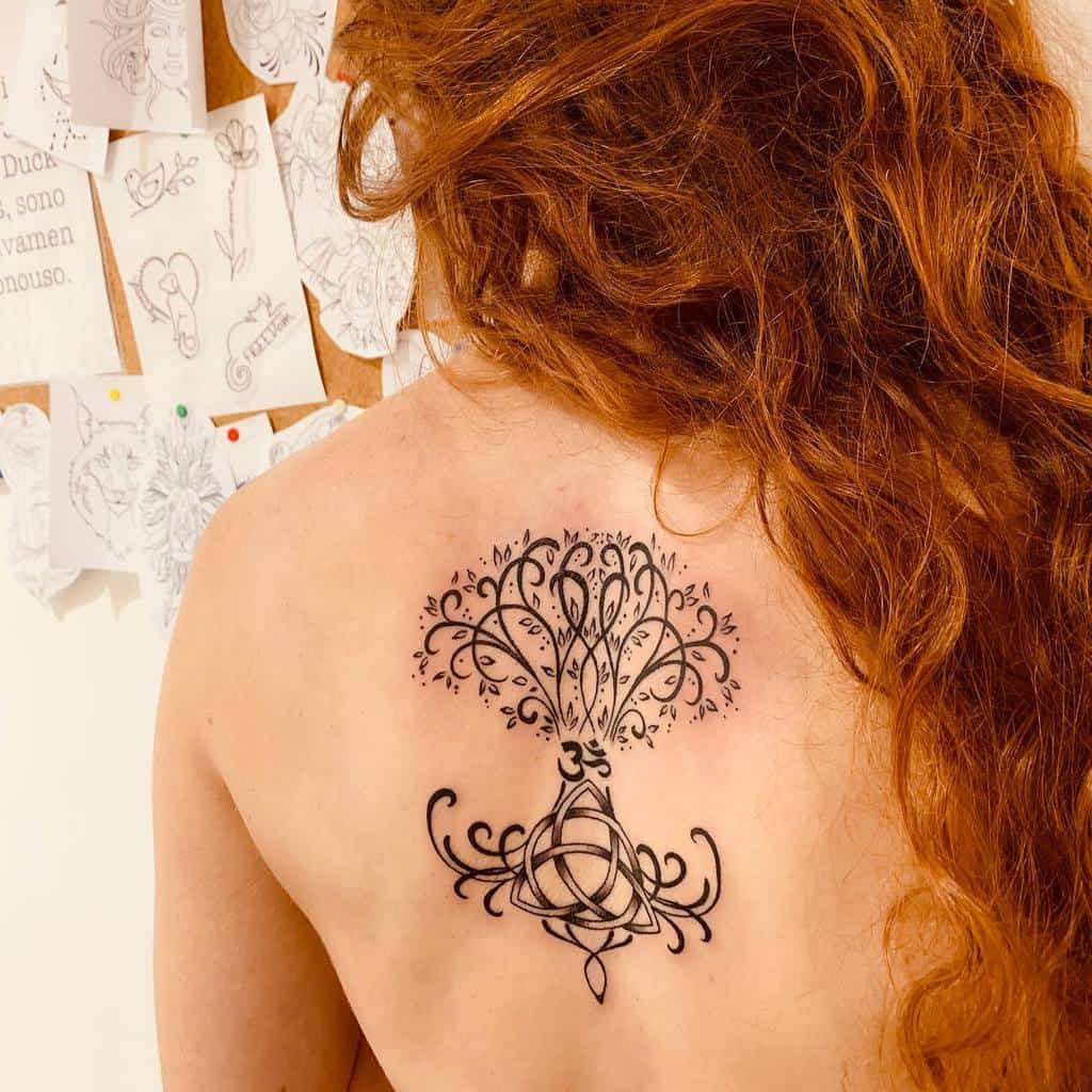 A woman with long red hair displays a black Celtic tattoo on her upper back, featuring a tree of life design with intricate knotwork and a triquetra at the base