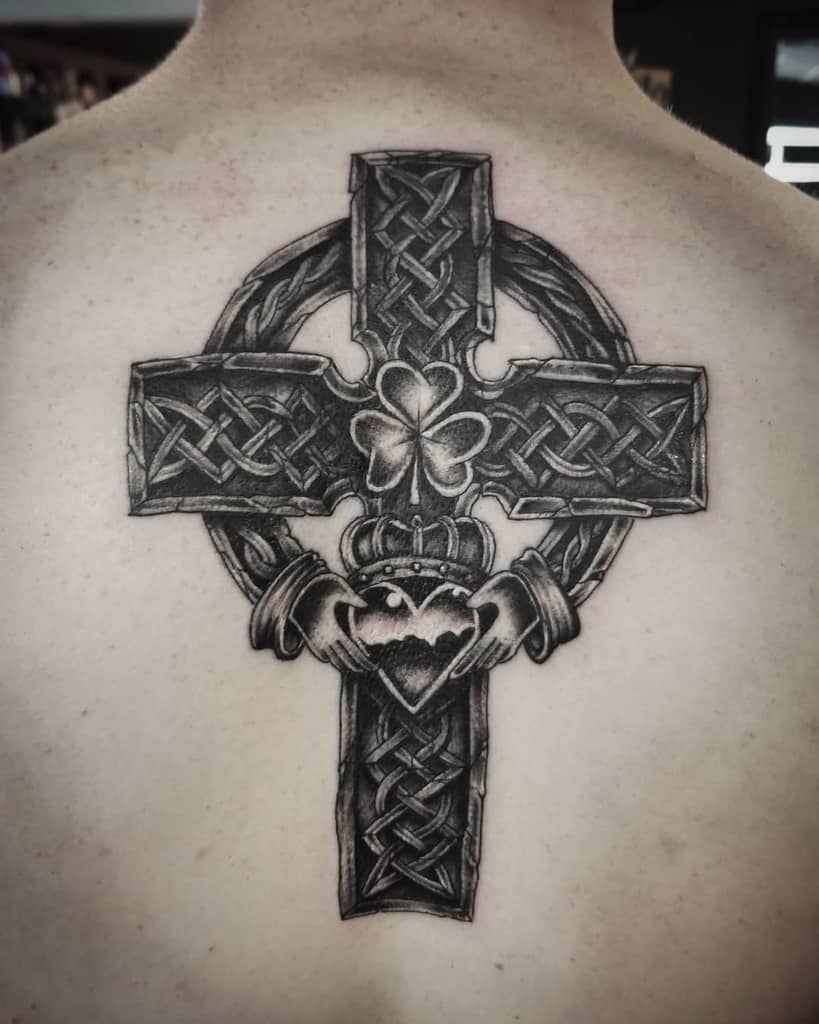 A Celtic cross tattoo elegantly adorns the person's back, intricately interwoven with a Claddagh symbol and a shamrock, capturing deep cultural significance and heritage