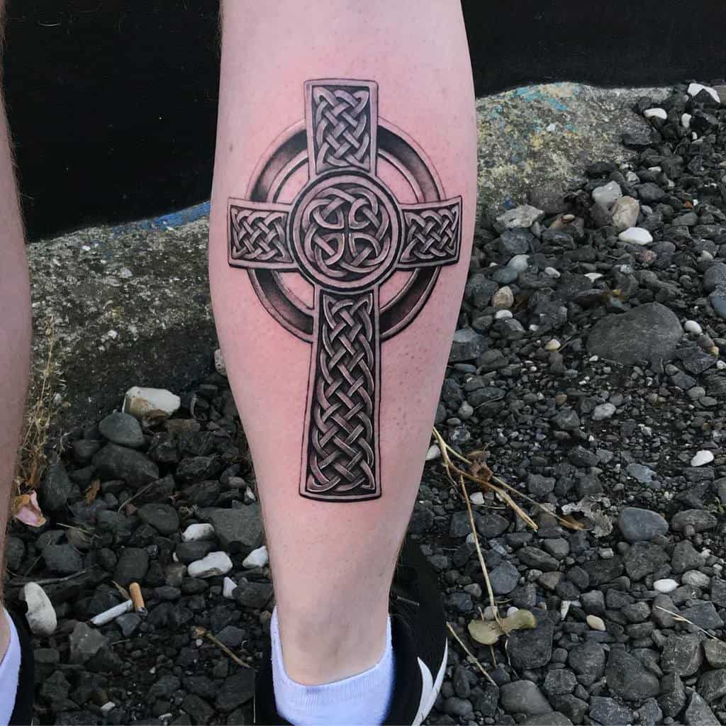 A Celtic cross tattoo featuring intricate knotwork graces a person's calf