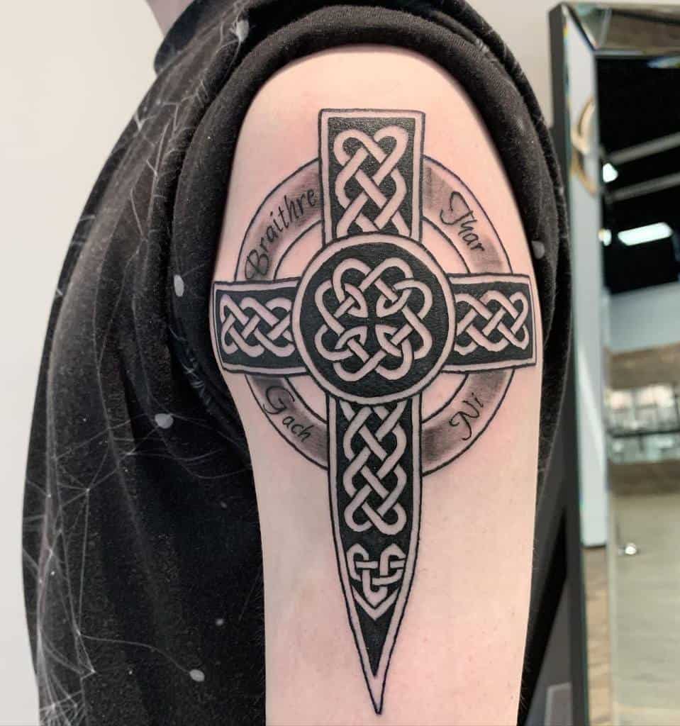 Celtic cross tattoo with intricate knot patterns and words on the upper arm, featuring a round design in the center