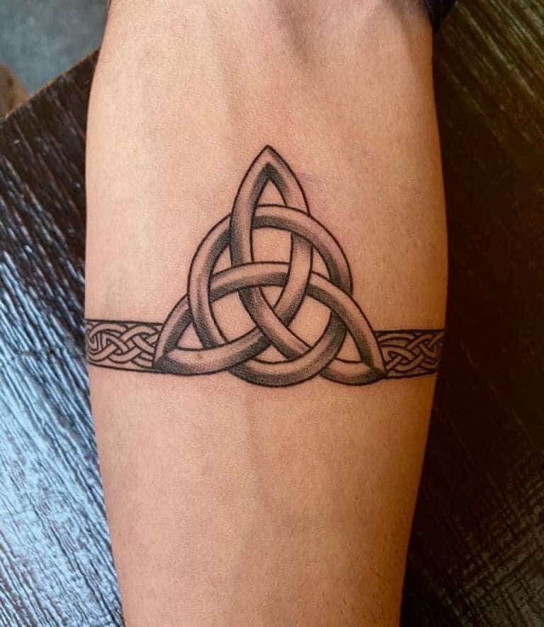 A Celtic knot tattoo on the forearm featuring an intricate triquetra design with a continuous band of knotwork encircling the arm