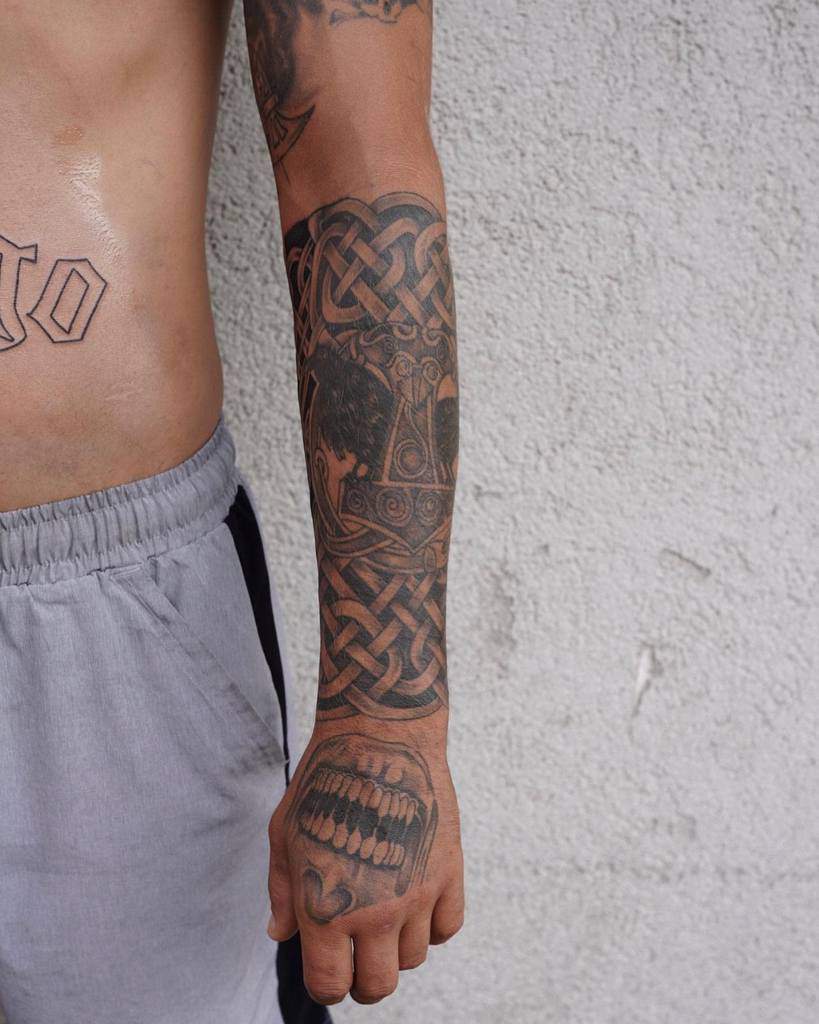 A Celtic-inspired forearm tattoo featuring intricate knotwork, Thor's hammer, and a hand tattoo with a bold set of fangs