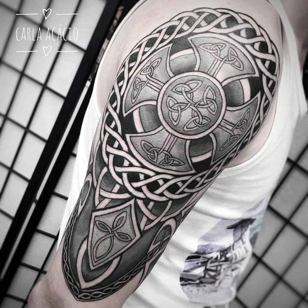A detailed Celtic shoulder tattoo featuring intricate knotwork, a central Celtic cross with triquetra symbols, and bold shading