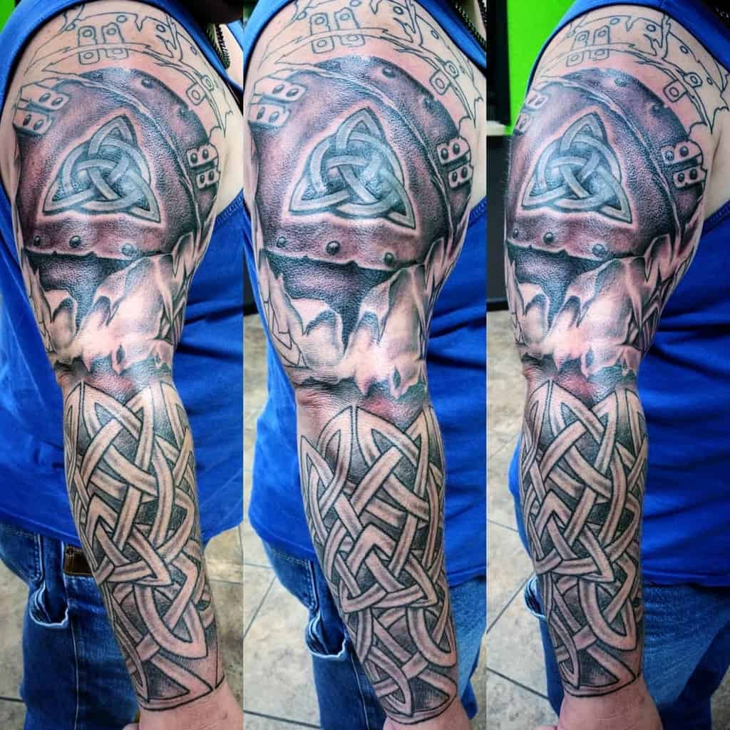 A full sleeve Celtic tribal tattoo with a bold design featuring a prominent Triquetra symbol at the shoulder encased in a 3D armor effect