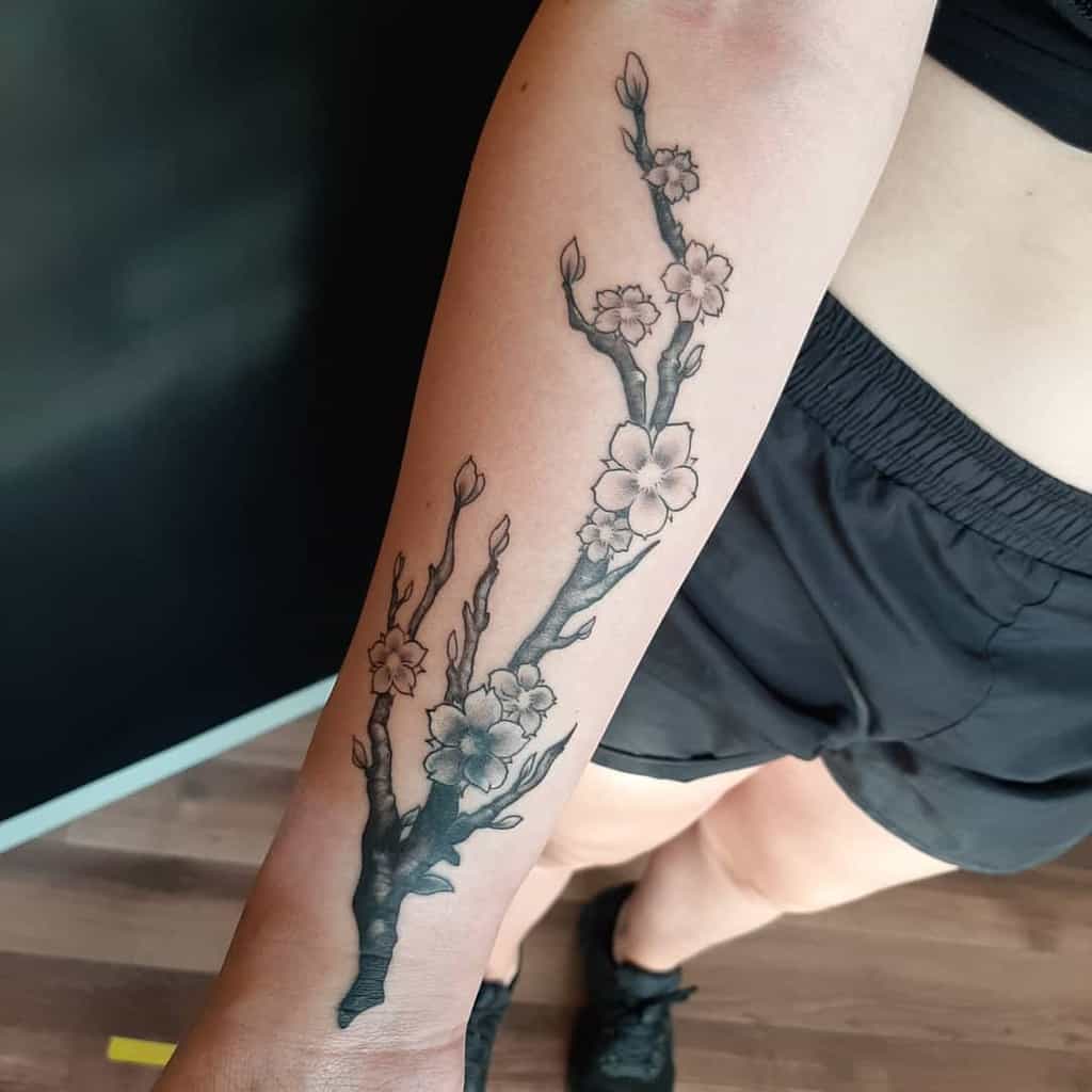 Forearm tattoo featuring a graceful branch intertwined with blooming flowers and buds, drawn in black and grey ink