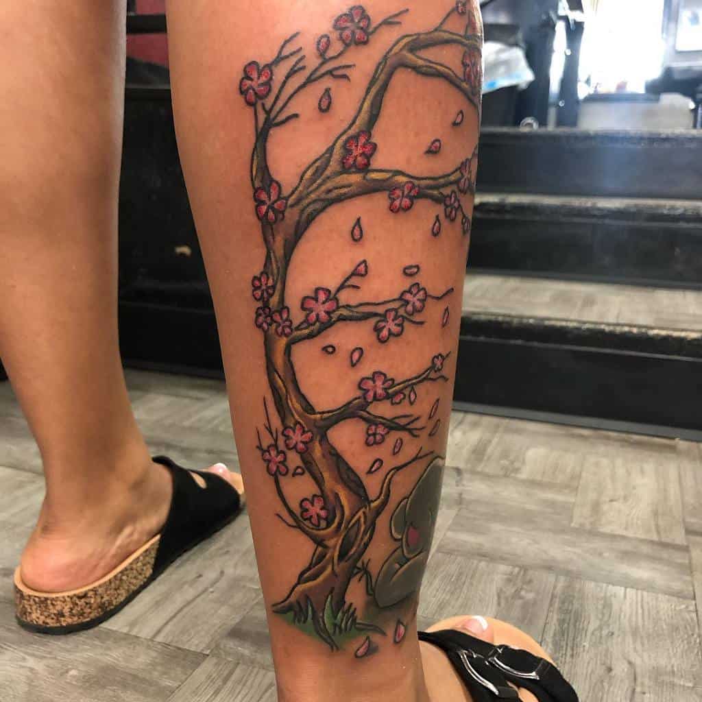 Leg tattoo of a cherry blossom tree with pink flowers and falling petals, featuring an intricate branch design on the back of a person's calf