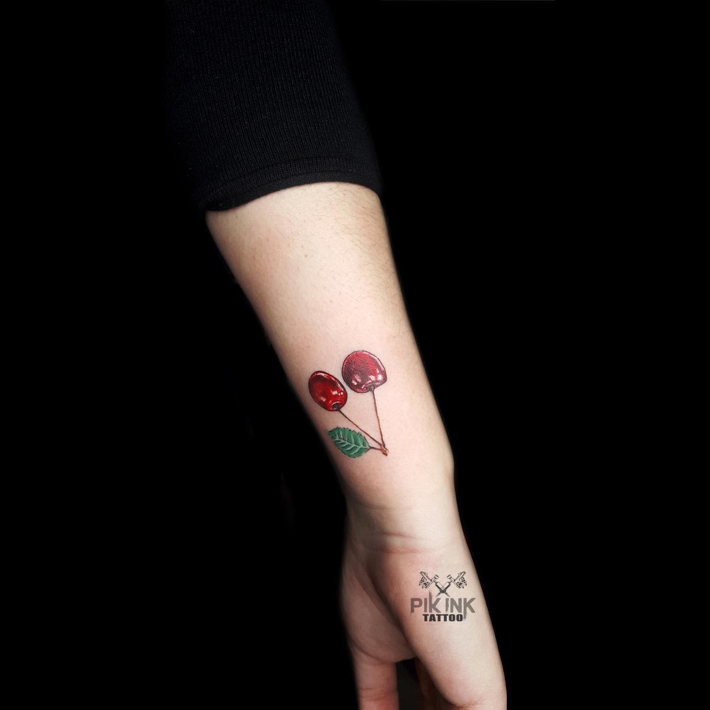 A tattoo of two red cherries with a green leaf on a forearm
