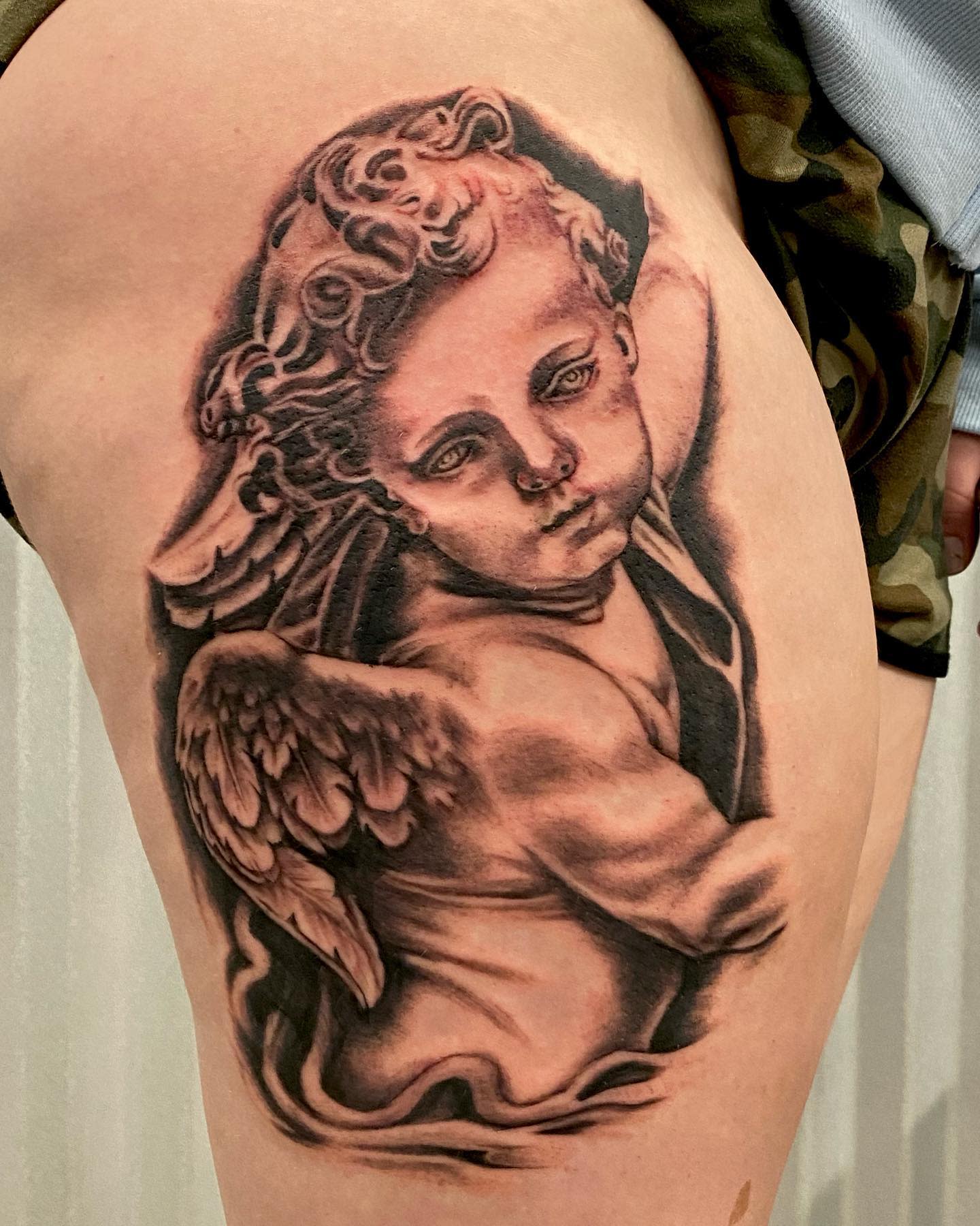 Tattoo of a detailed cherub with wings on a thigh, reminiscent of classical art