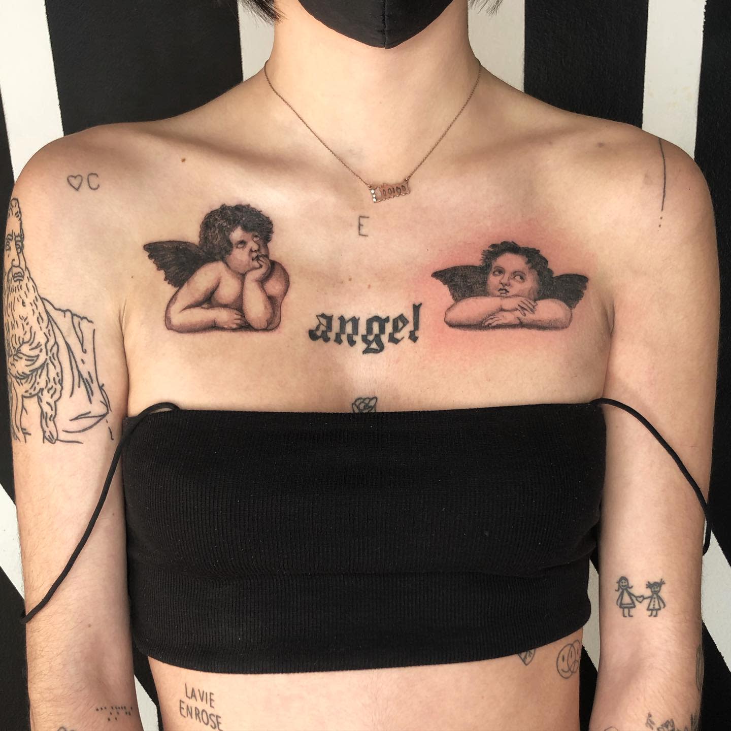 Cherub tattoos on shoulders and "angel" text tattoo on chest