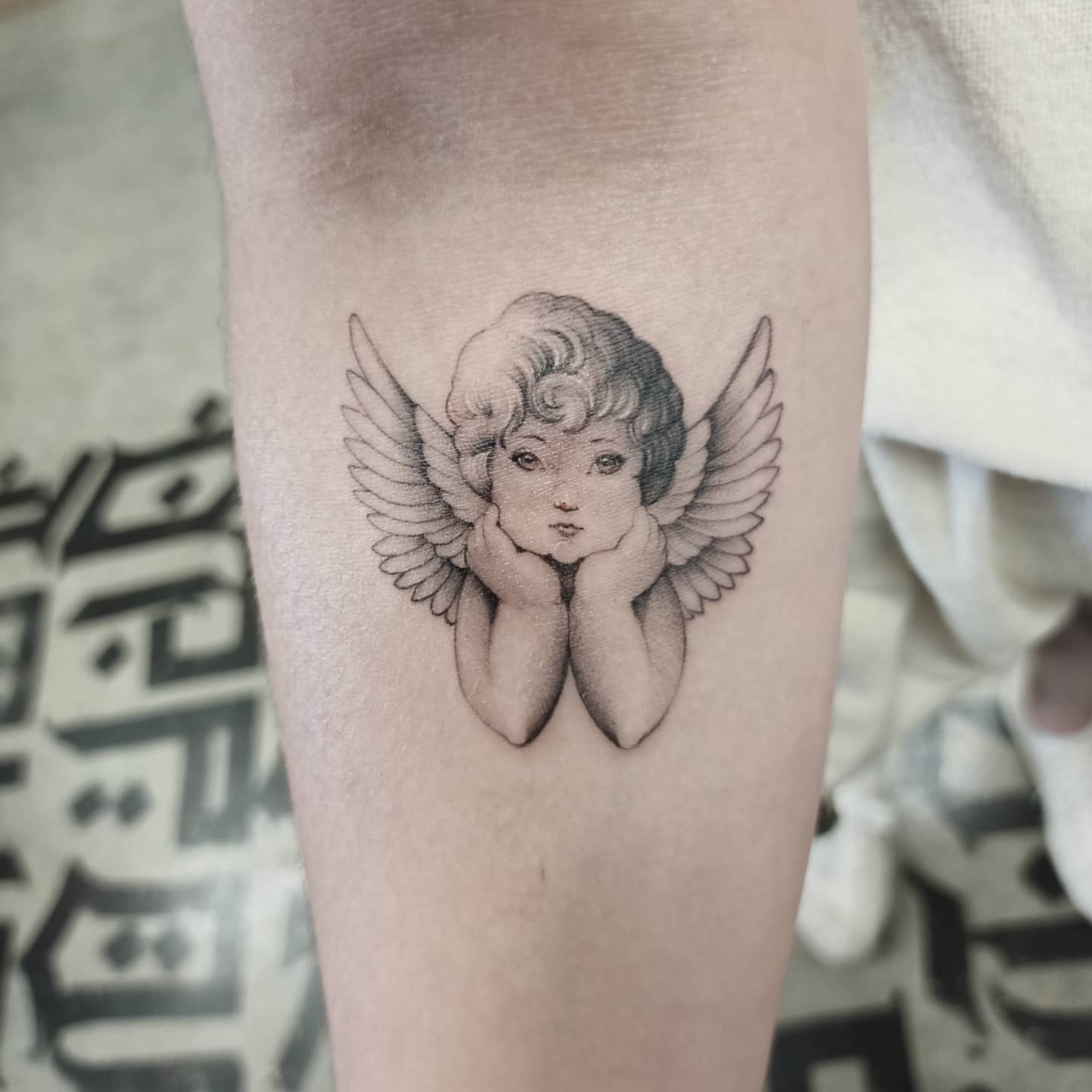 Tattoo of a cherub with wings resting its chin on its hands, inked on an arm