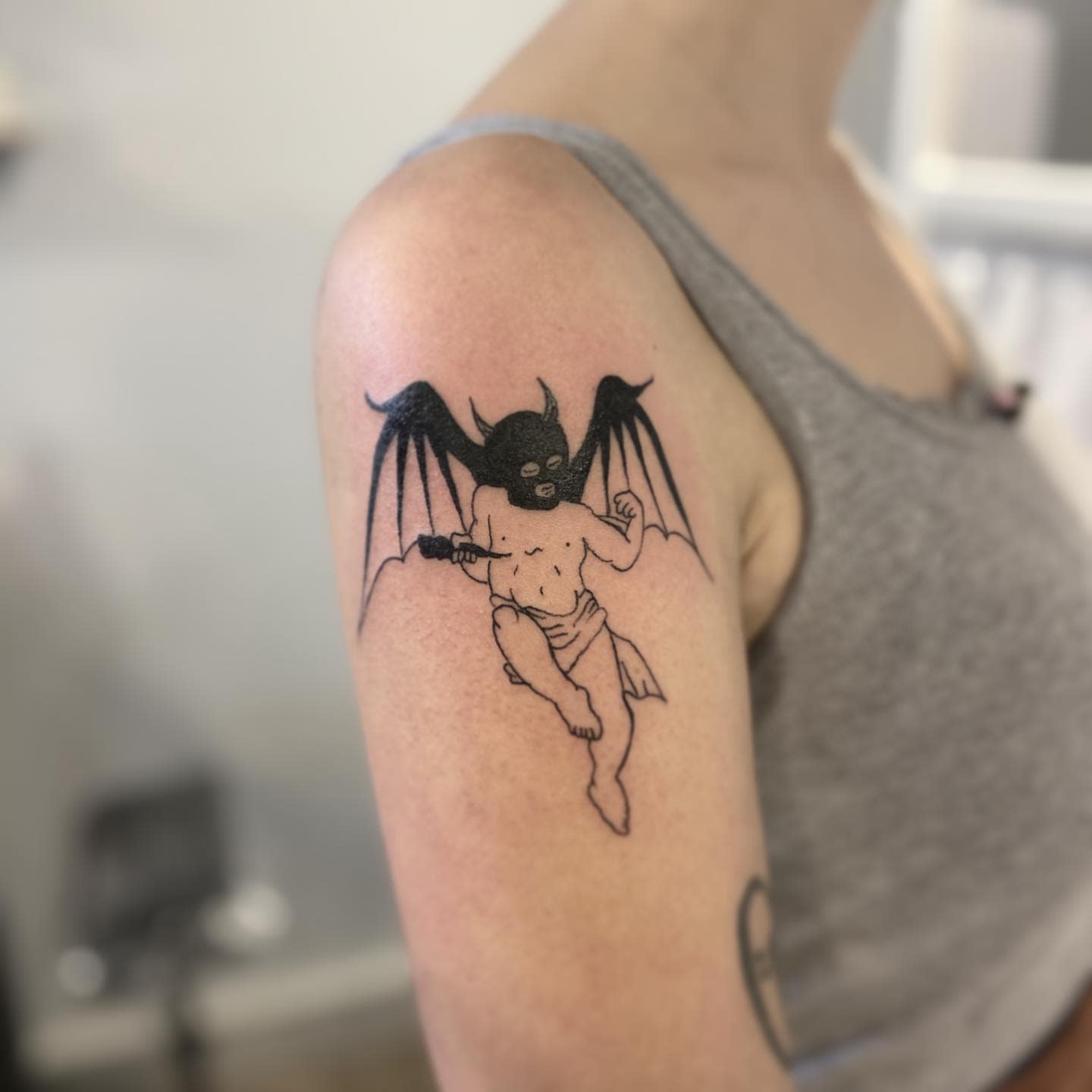 Tattoo on upper arm of an evil cherub wearing a dark mask and holding a stick