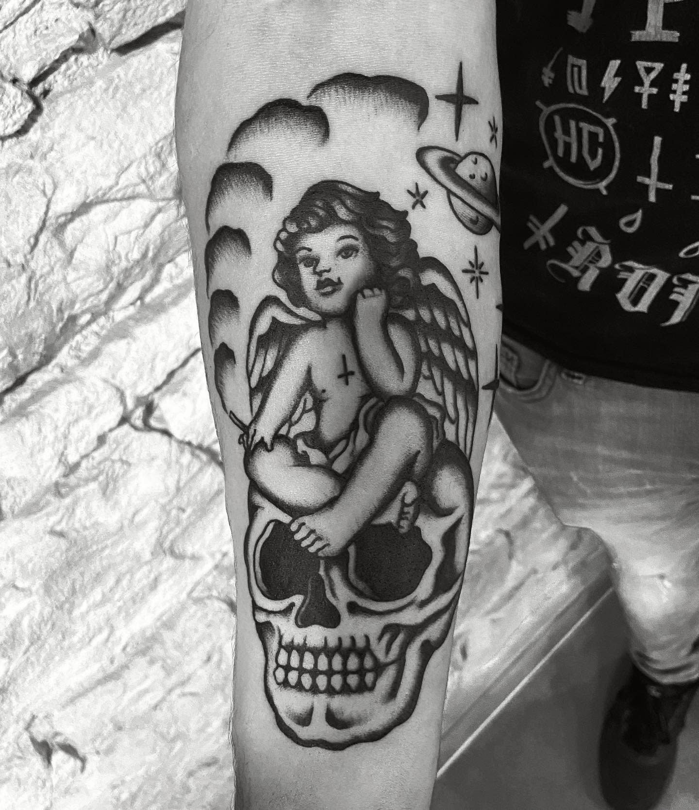 Tattoo depicting a cherub atop a skull, set against a backdrop of space and religious icons