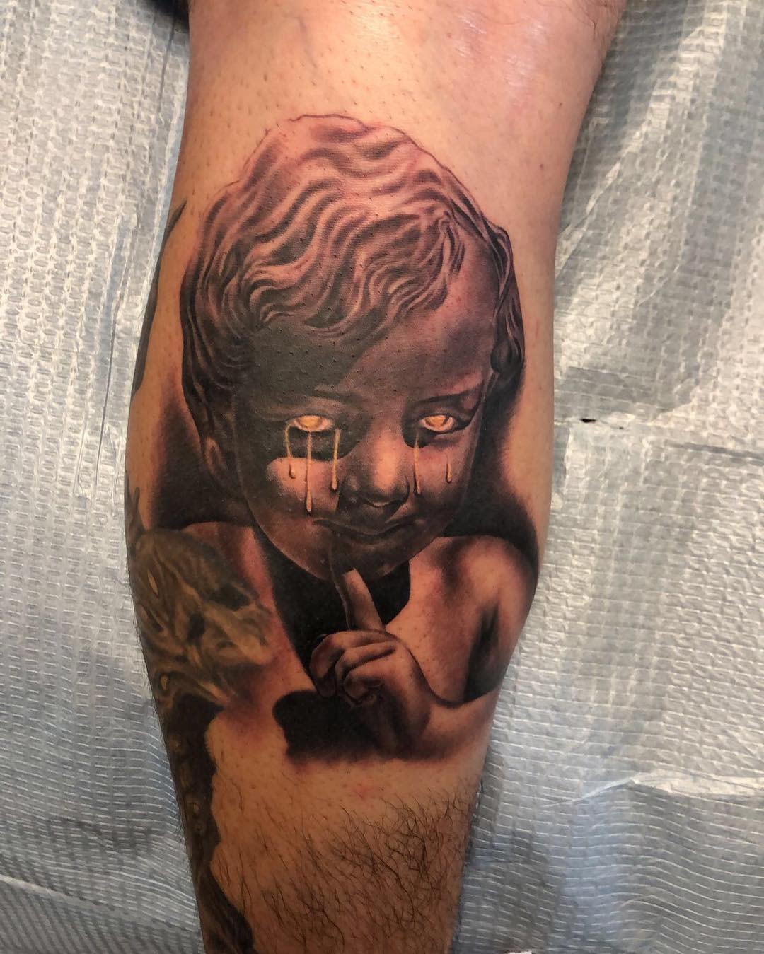 Realistic tattoo of a crying cherub's face with finger on lips, located on leg
