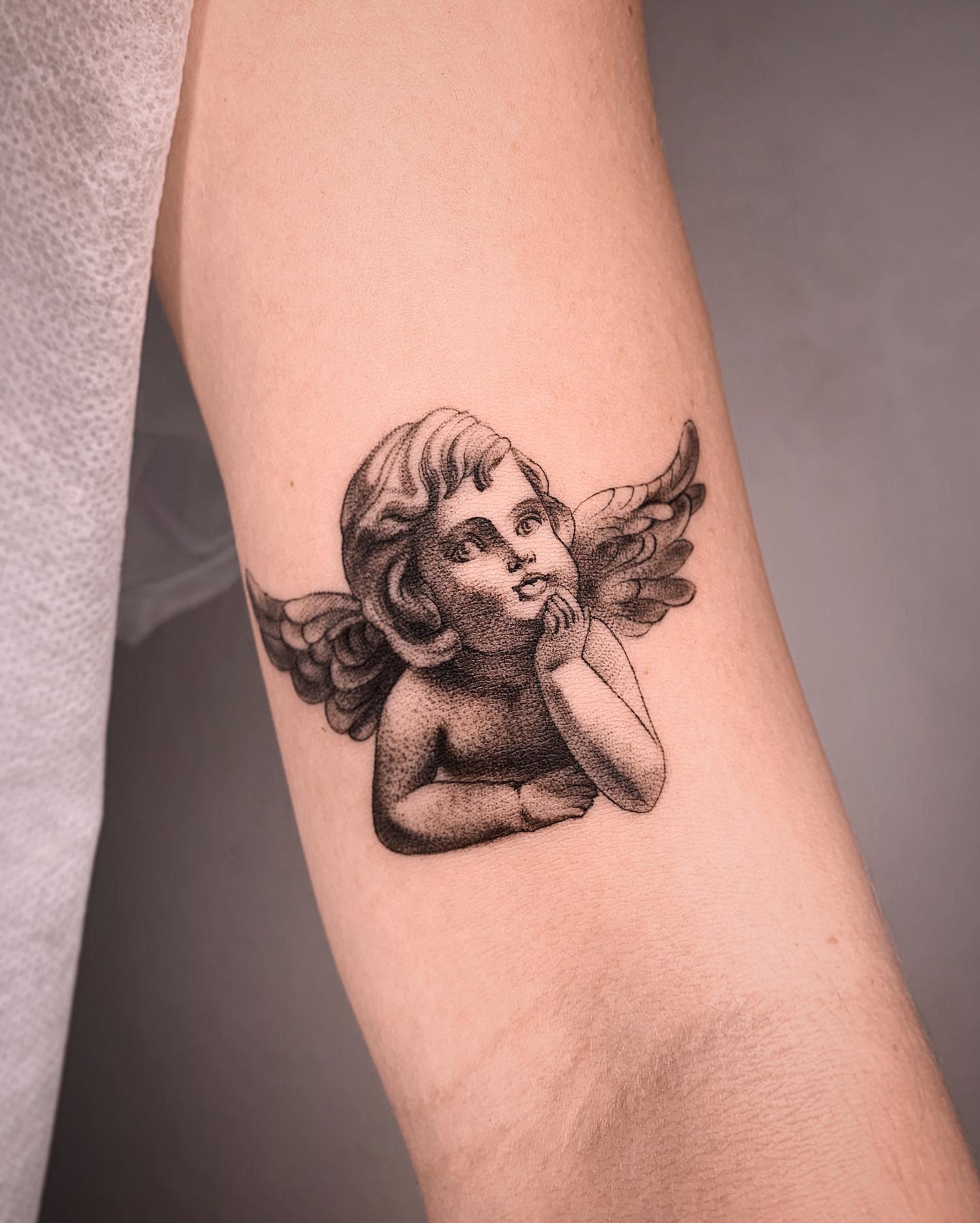 Tattoo of a cherub with wings, chin resting on hand, on the inside of an arm
