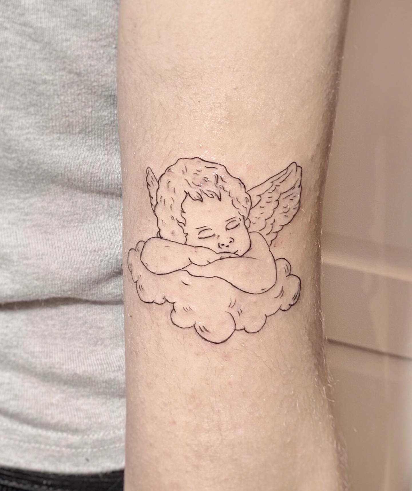 Line tattoo of a sleeping cherub with wings, resting on a cloud, on an inner forearm