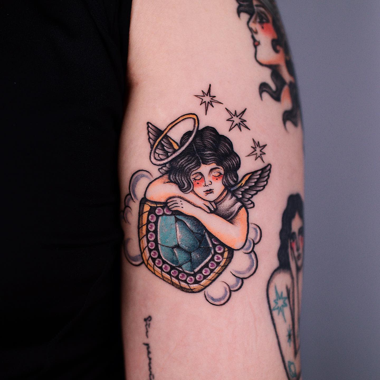 Tattoo of a sad cherub with wings and a halo, resting on a gem with stars and clouds