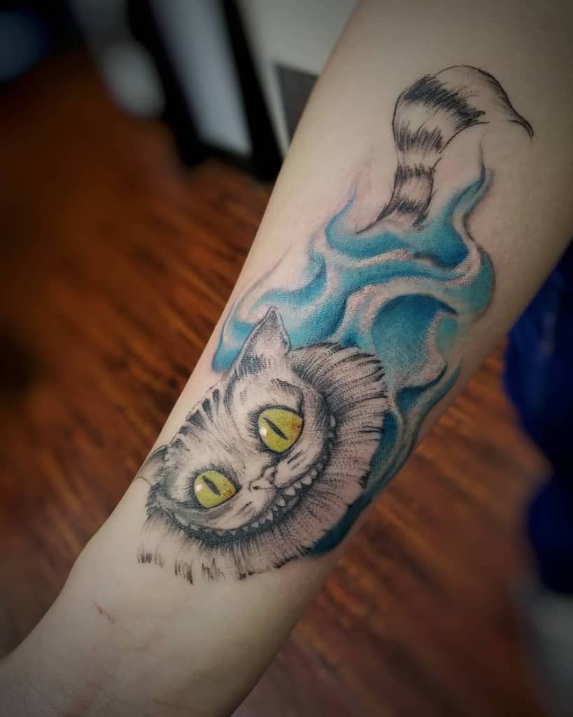 A Cheshire Cat forearm tattoo with glowing yellow eyes, floating in blue smoke, showcasing its signature grin