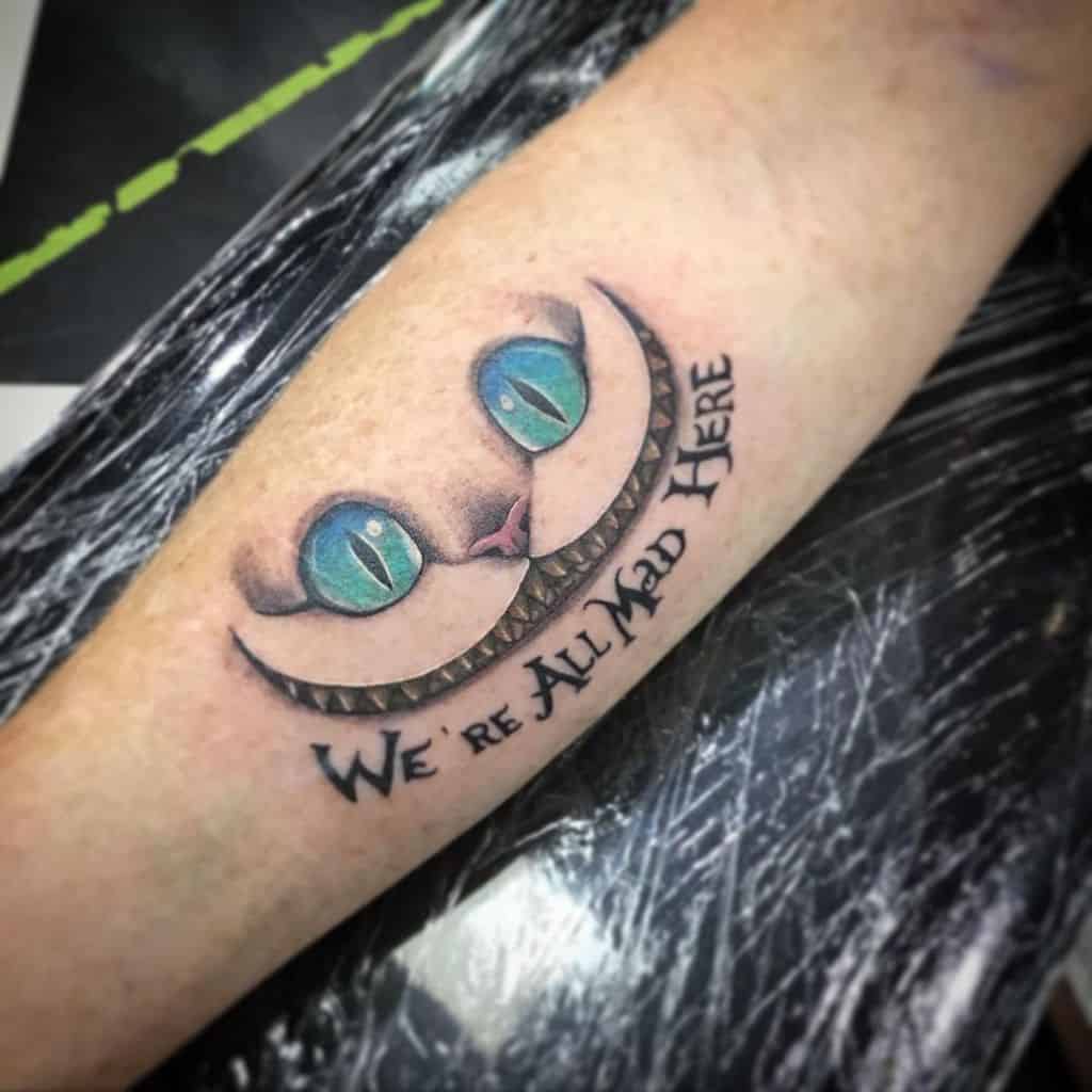 A Cheshire Cat tattoo featuring its eyes and wide grin, with the quote "We're all mad here" curved below