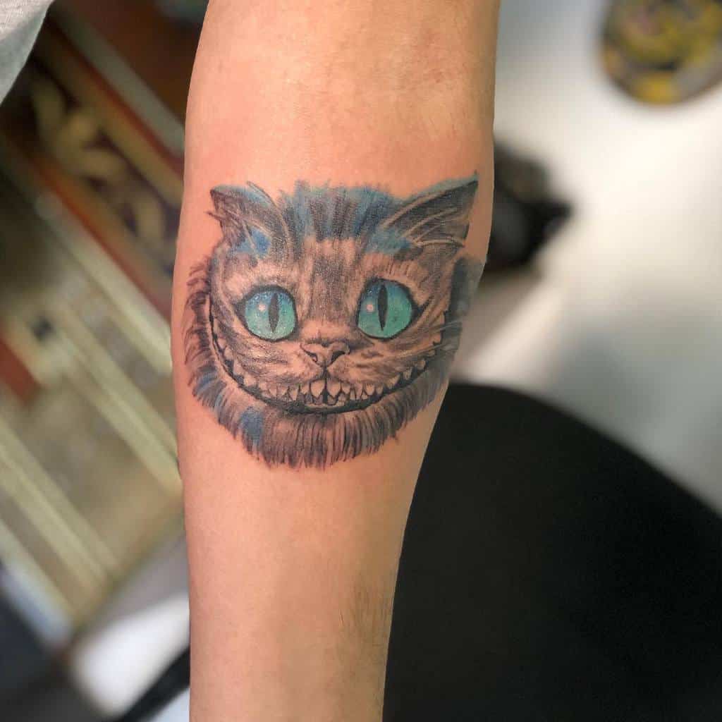 A Cheshire Cat forearm tattoo featuring its iconic wide grin and glowing blue eyes, set against subtle shading