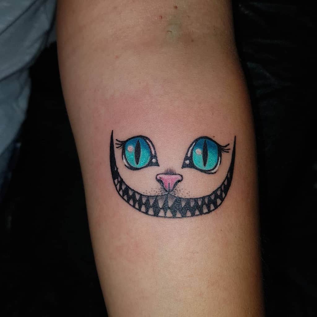 A minimalist Cheshire Cat tattoo featuring its signature wide grin, bright blue eyes, and a small pink nose