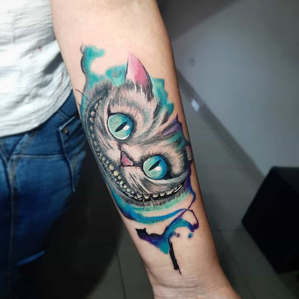 A vibrant Cheshire Cat forearm tattoo with glowing blue eyes and a wide grin, surrounded by blue and purple smoky accents