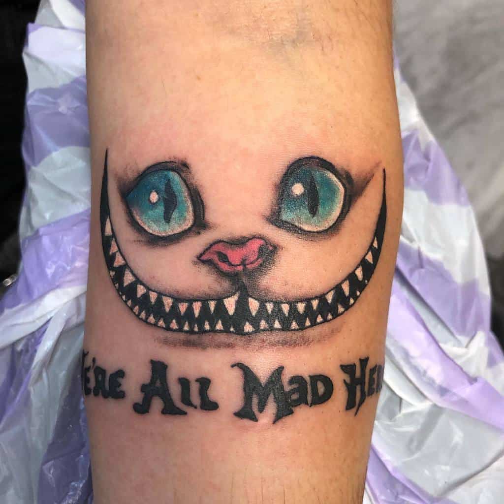 A tattoo featuring the Cheshire Cat’s eyes, wide grin, and the quote "We’re All Mad Here" beneath it