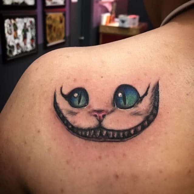 A tattoo on the shoulder featuring the Cheshire Cat’s wide grin and bright green eyes, capturing its mischievous expression