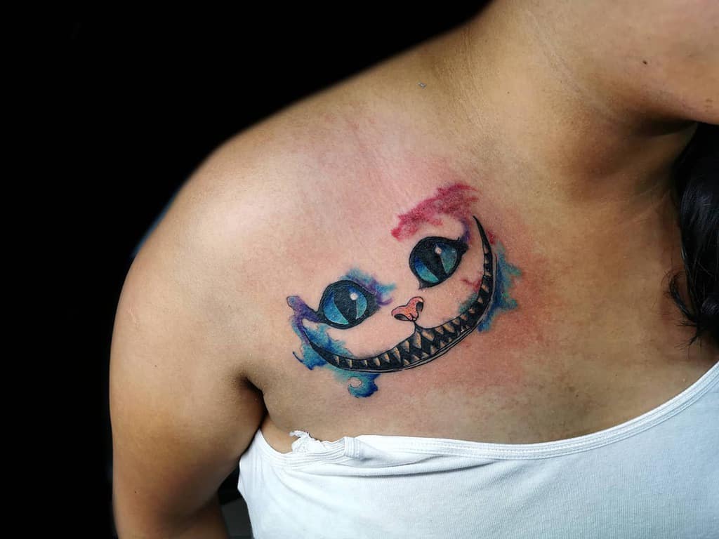 A watercolor-style tattoo on the chest featuring the Cheshire Cat’s eyes and wide grin with colorful accents in pink and blue