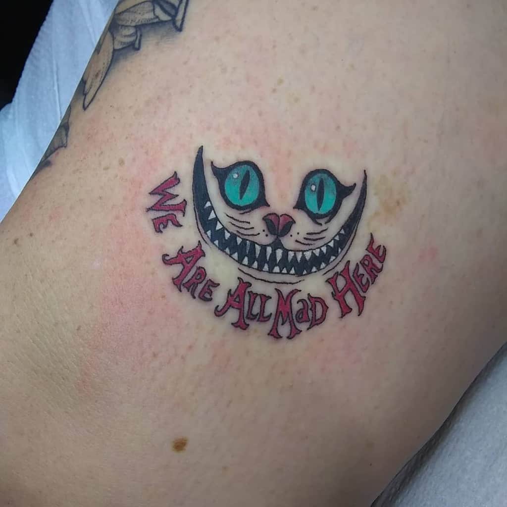 Tattoo of the mischievous Cheshire Cat's face with the text "We Are All Mad Here" beneath it
