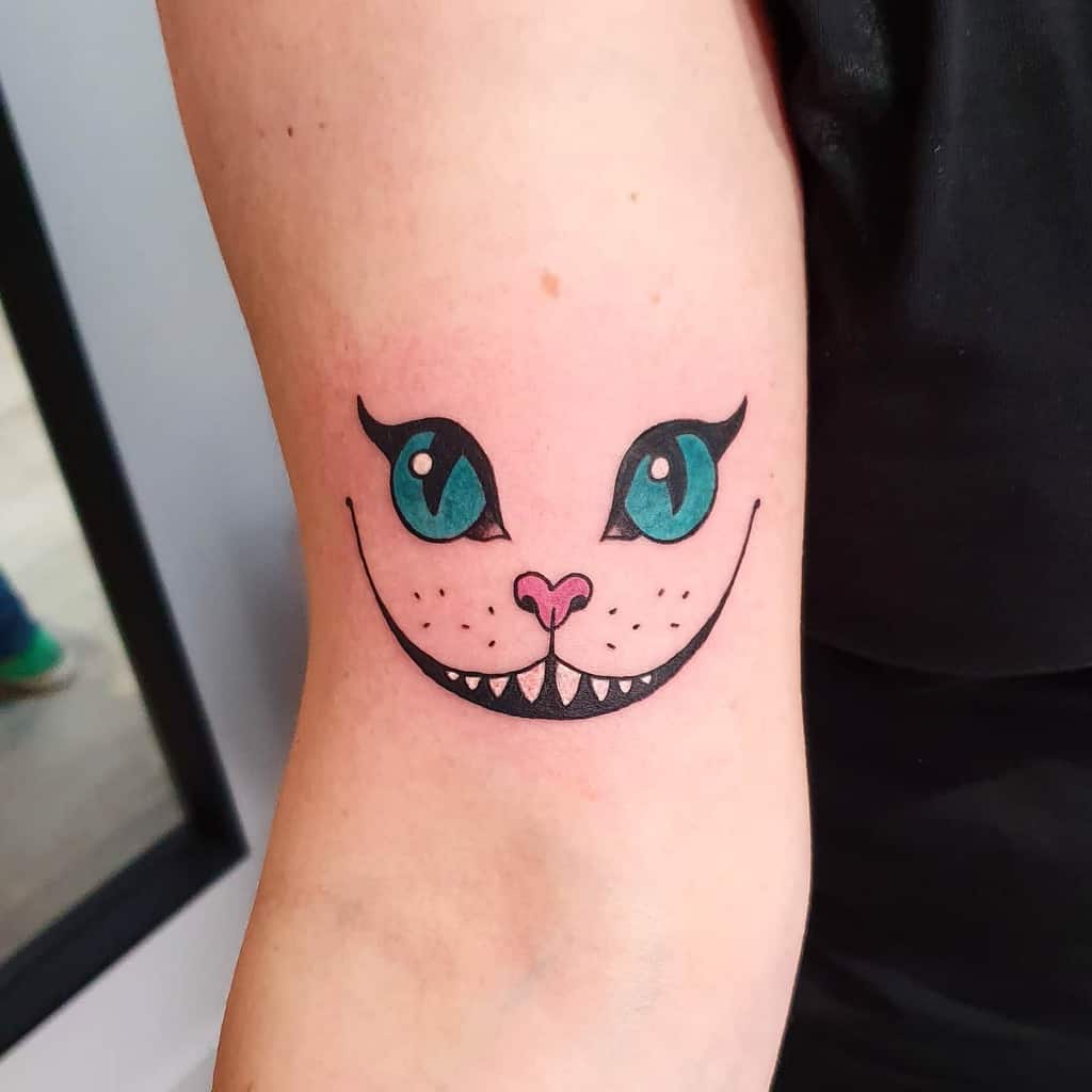 Tattoo of a Cheshire Cat face with big blue eyes and pointed teeth on a person's arm, exuding that signature mischievous grin