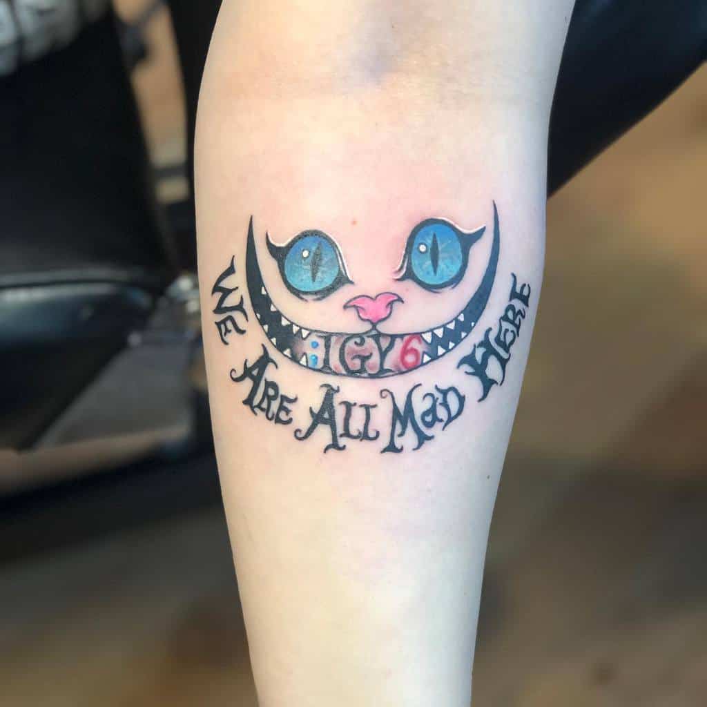Tattoo of Cheshire Cat's face with text "We Are All Mad Here" on a forearm