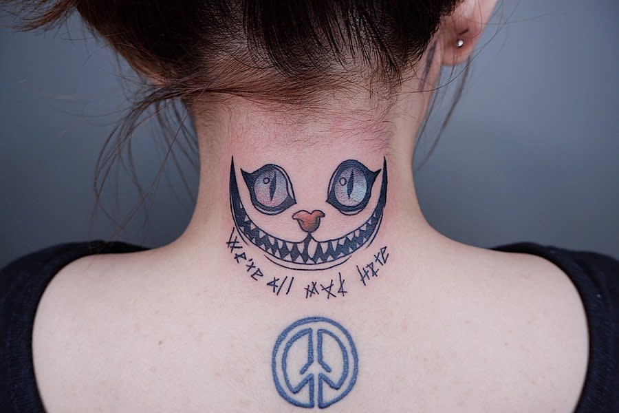 Tattoo of a grinning Cheshire cat face with eyes on the neck and "We're all mad here" text below