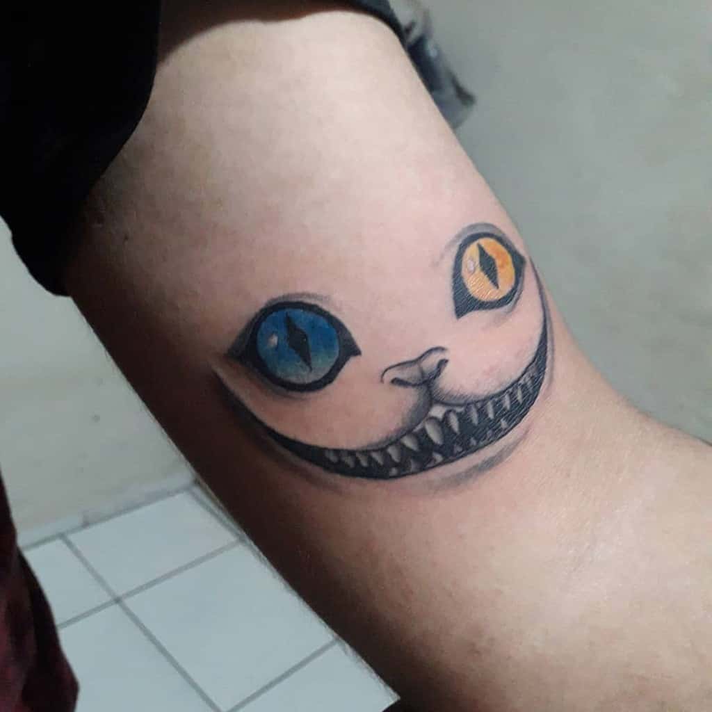 Tattoo of a grinning Cheshire cat with one blue eye and one yellow eye on an arm
