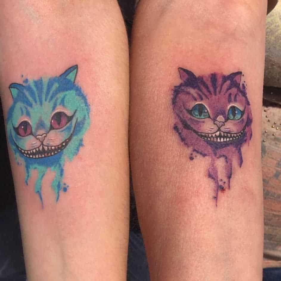 Colorful tattoos of the Cheshire Cat from Alice in Wonderland, with one tattoo in blue and the other in purple, with watercolor-like splashes
