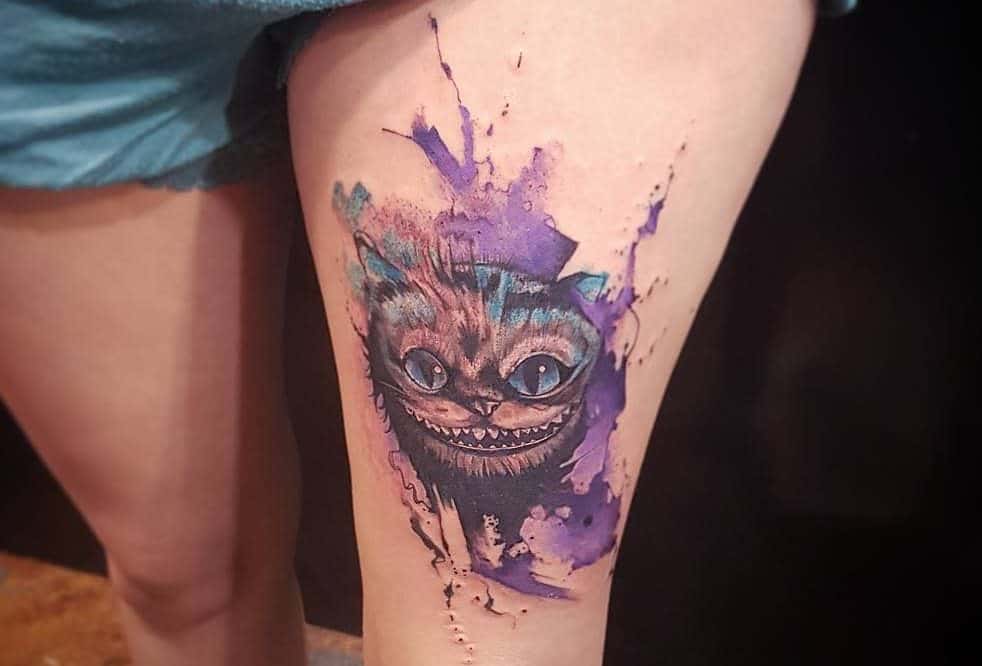 Vibrant tattoo of the Cheshire Cat from Alice in Wonderland, surrounded by colorful purple and blue watercolor splashes
