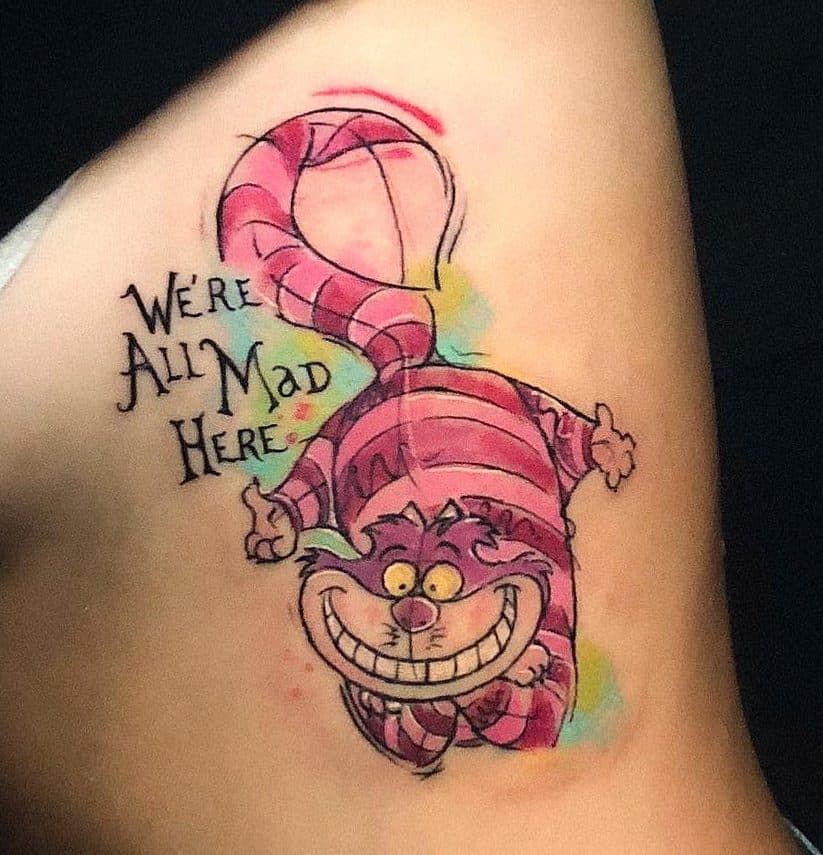 Colorful tattoo of the Cheshire Cat from Alice in Wonderland, with the quote 'We're all mad here', surrounded by splashes of pink, green, and yellow watercolor