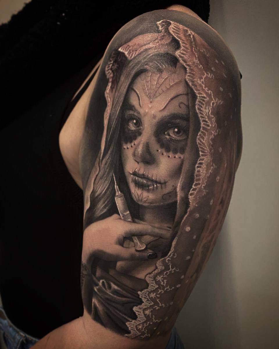 A detailed Catrina tattoo featuring a woman with sugar skull makeup, holding a syringe, draped in lace