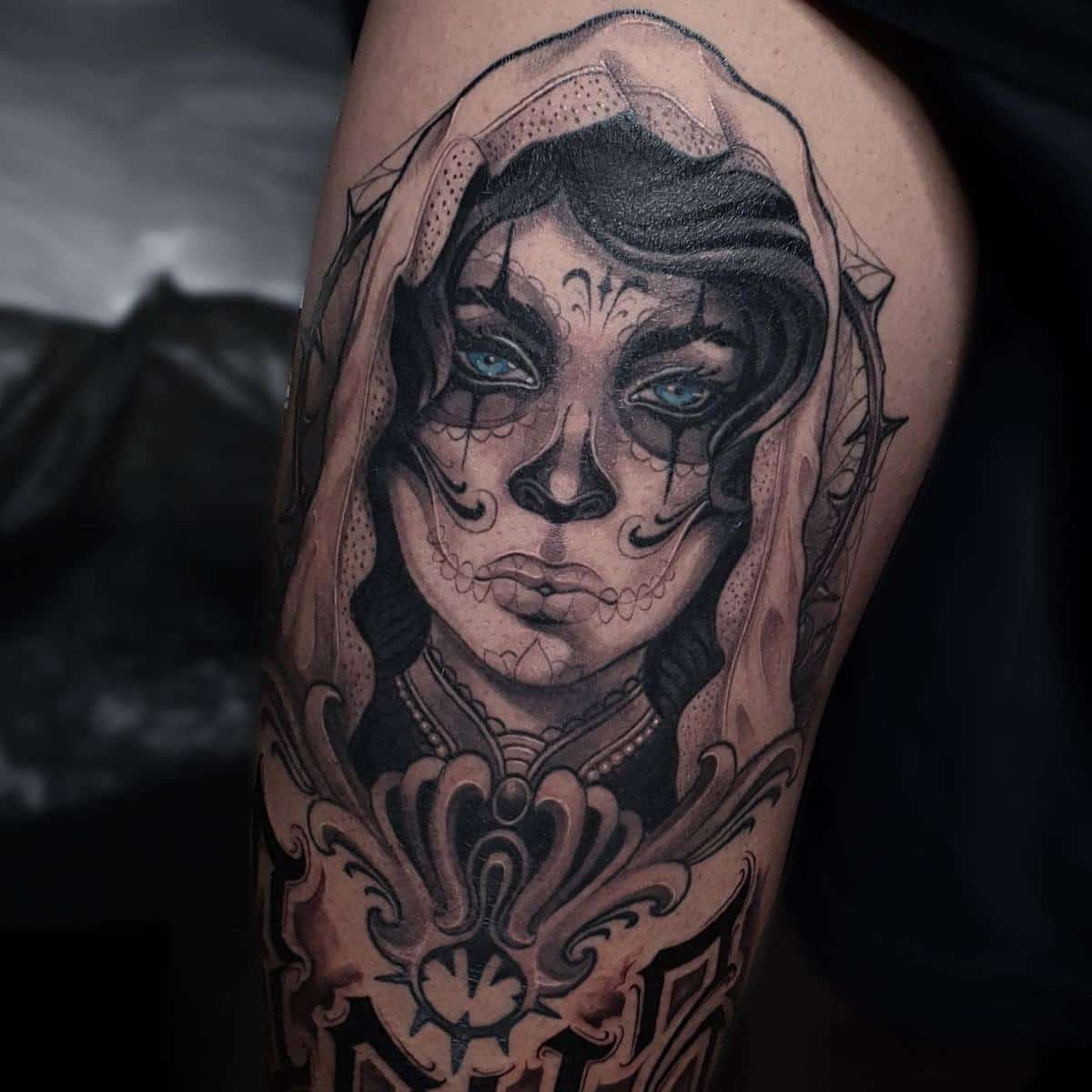 Detailed black-and-grey tattoo of a Catrina figure, with intricate face paint and vivid blue eyes, framed by ornate patterns