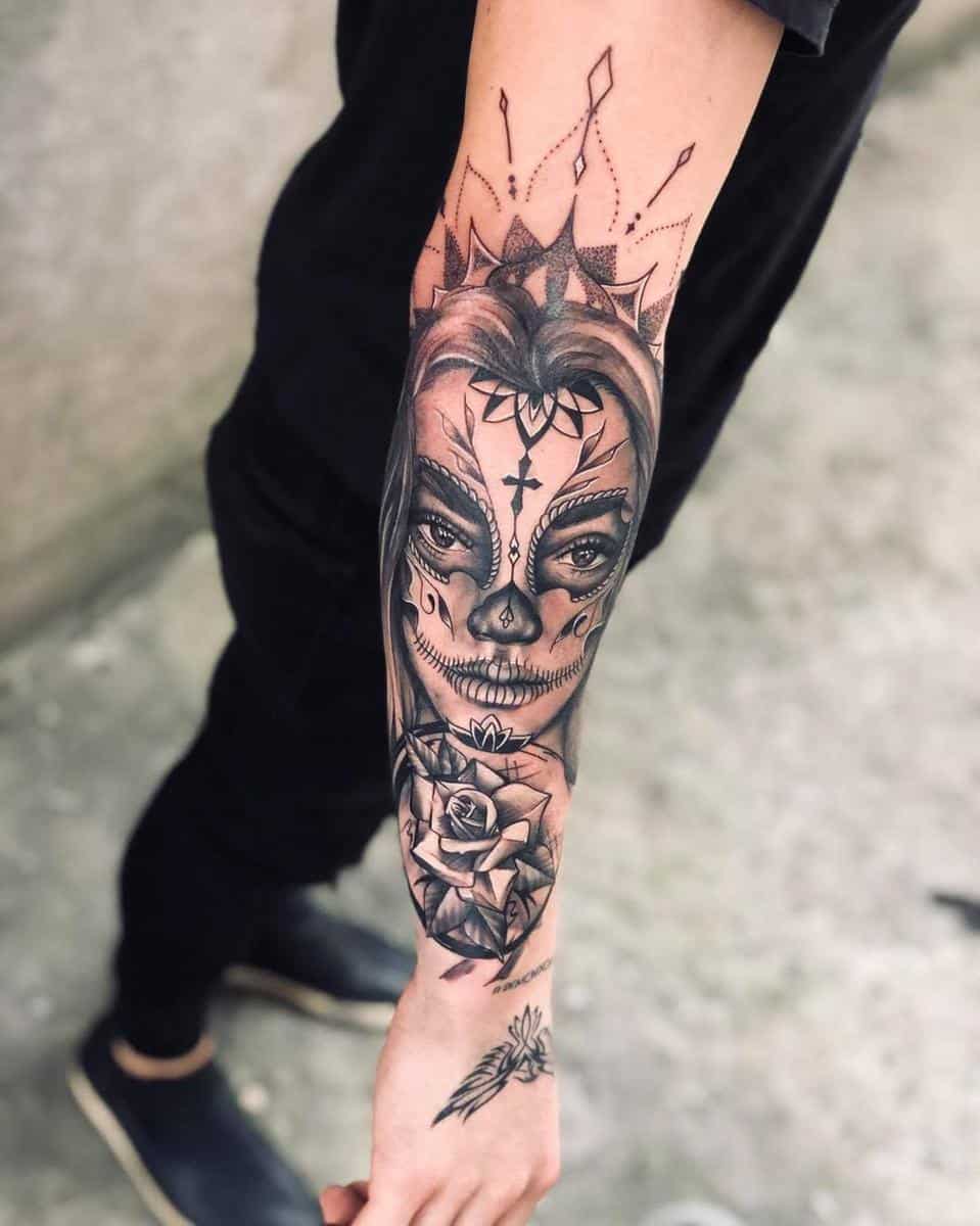 A Catrina tattoo on the forearm featuring a woman with sugar skull makeup, a crown of geometric shapes, and a rose