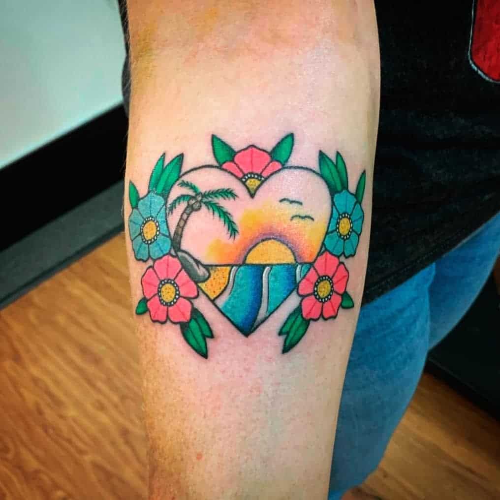 Tattoo of a heart with a sunset, palm tree, ocean, sand, rocks and surrounded by flowers with leaves