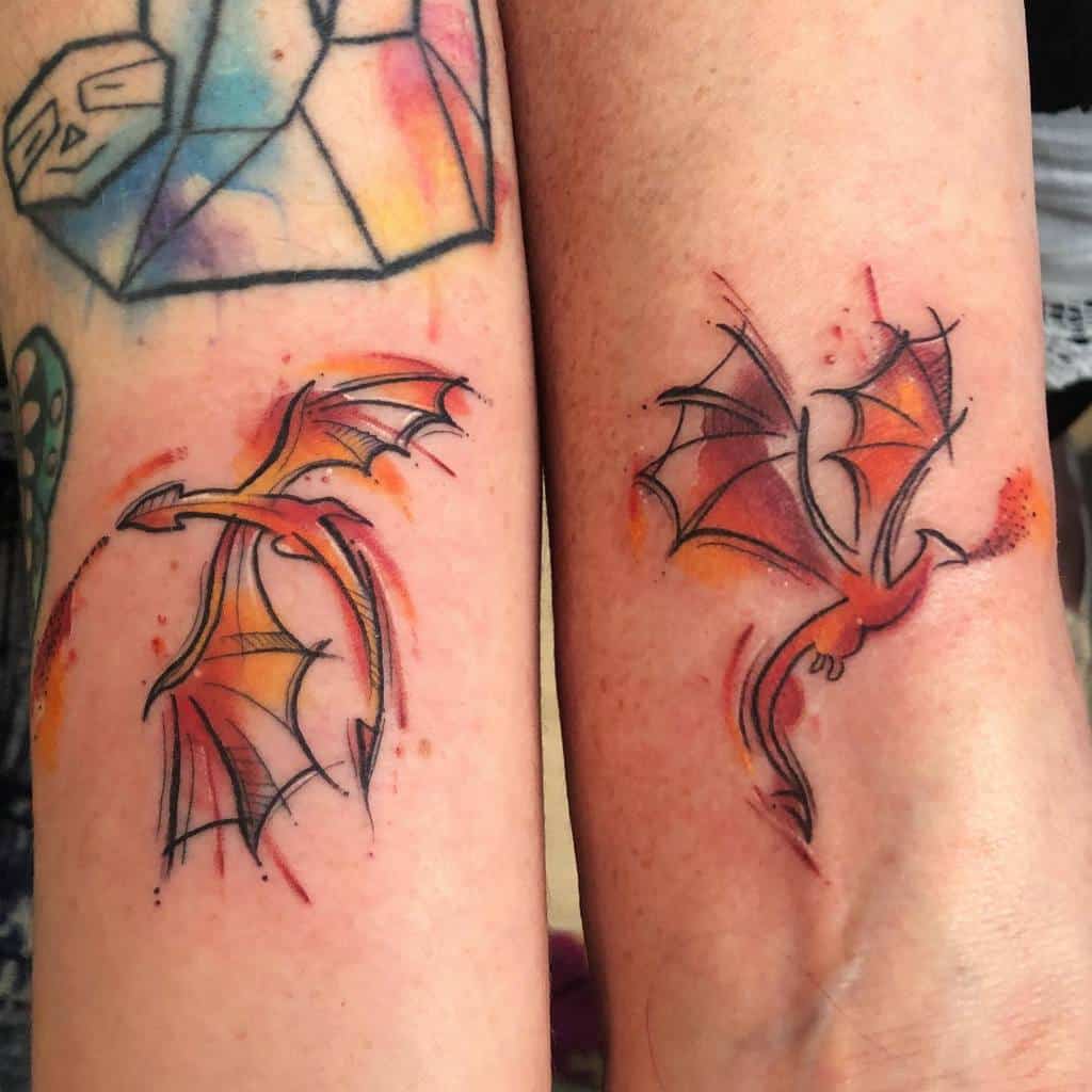Colorful watercolor-style tattoo of two small dragons in flight, with orange and red hues