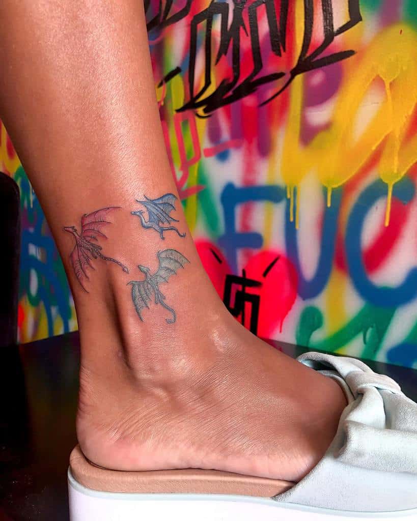 Ankle tattoo featuring three small, colorful dragons in flight, with pastel shades of pink, blue, and green
