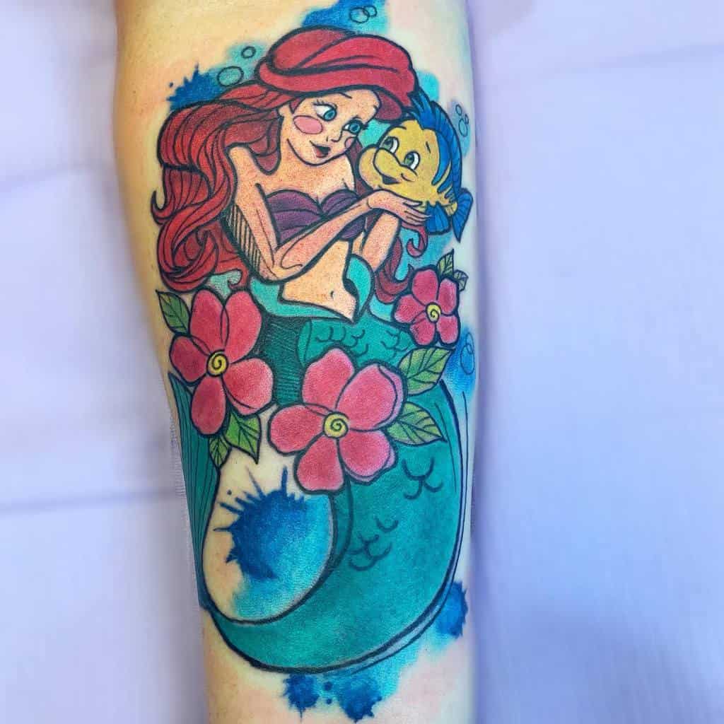 A vibrant tattoo of Ariel holding Flounder, surrounded by pink flowers, blue water splashes, and her iconic red hair and green tail