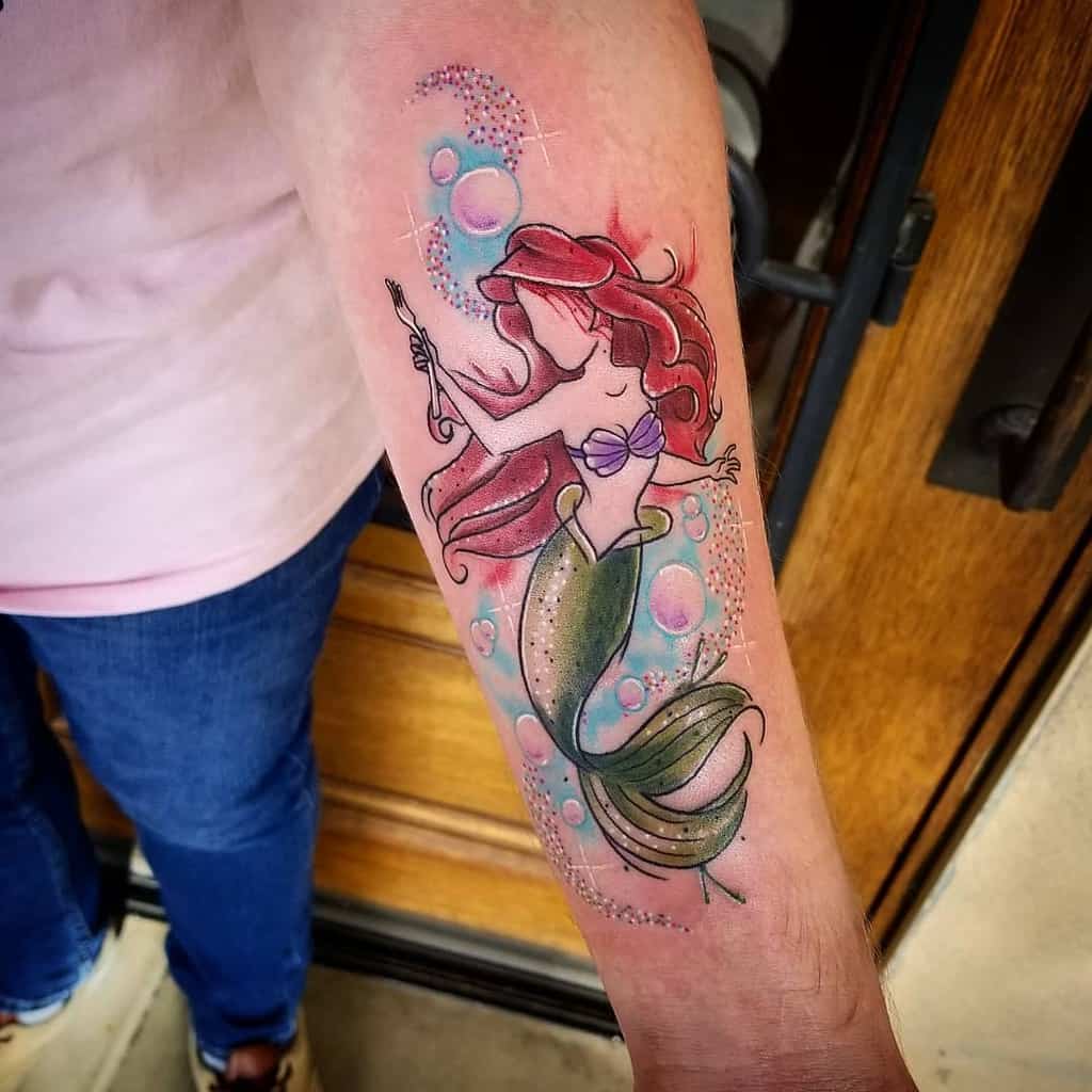 A whimsical tattoo of Ariel holding a fork, surrounded by sparkling bubbles, colorful accents, and her flowing red hair and green tail