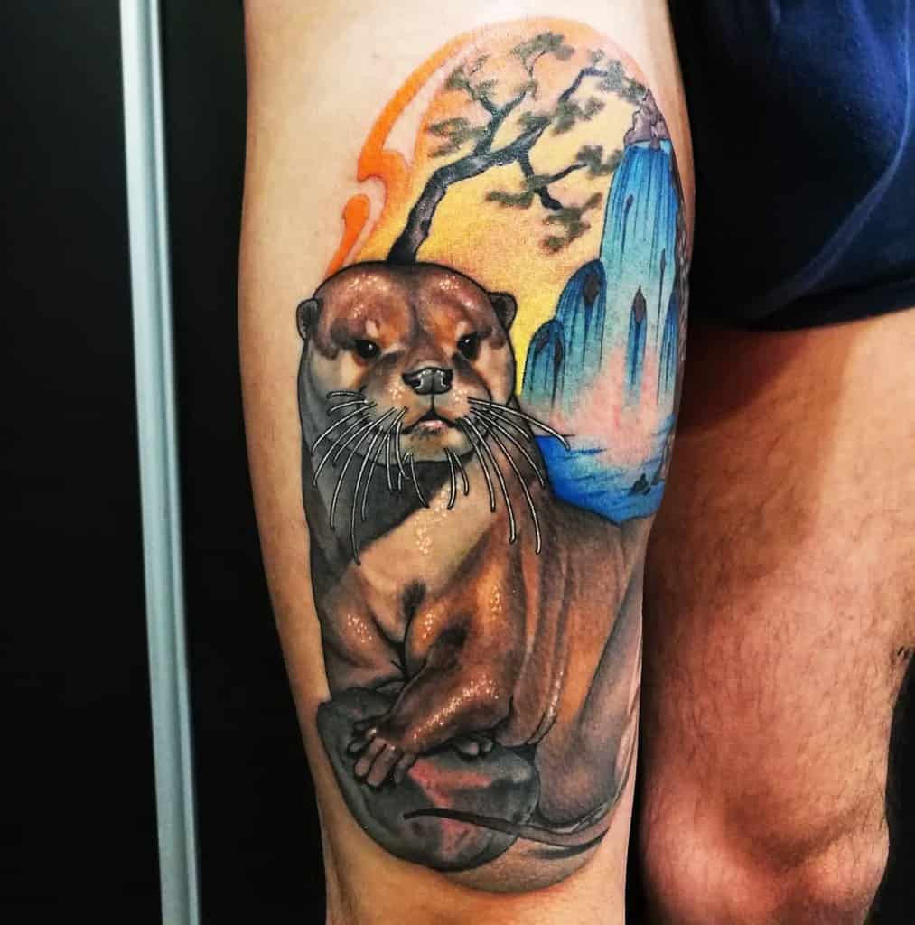 Otter tattoo with a vibrant tree and cliffs backdrop adorns the arm