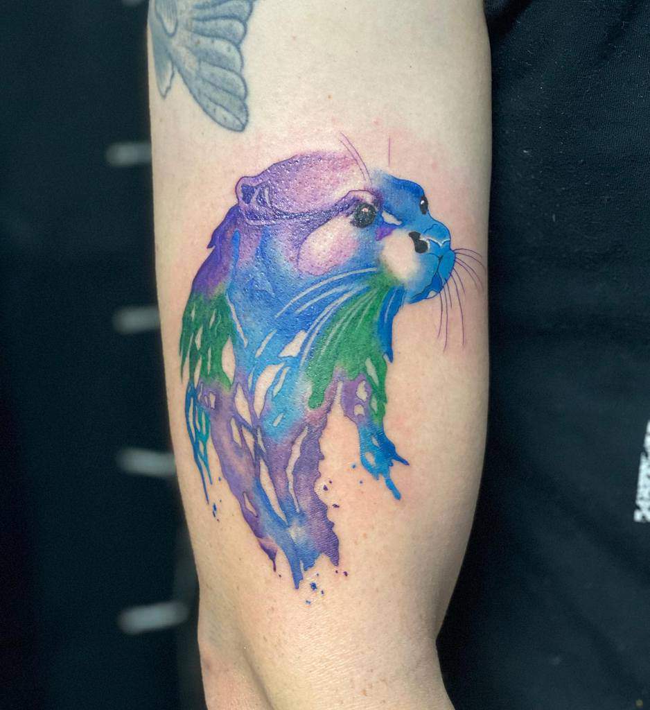 A colorful otter tattoo with a vibrant watercolor effect graces the arm, showcasing blues, purples, and greens