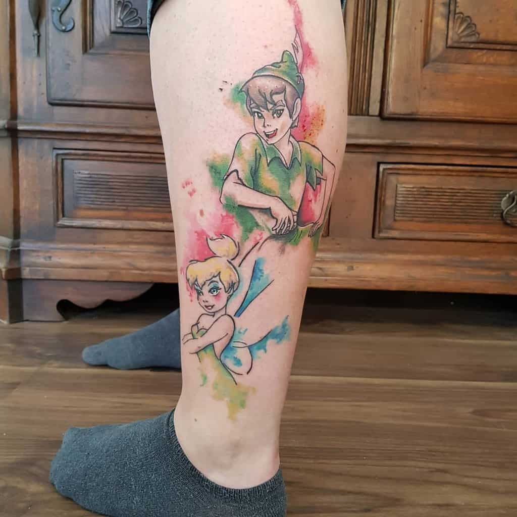 Watercolor-style tattoo of Peter Pan and Tinker Bell on a person's leg, featuring vibrant splashes of green, blue, red, and yellow