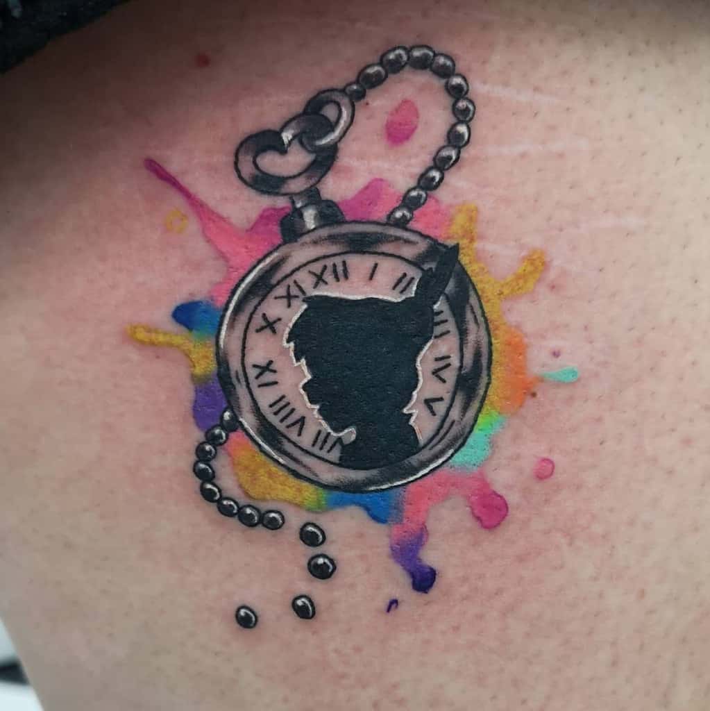Black and gray pocket watch tattoo with Peter Pan’s silhouette, set against a vibrant watercolor splash of pink, yellow, blue, and purple